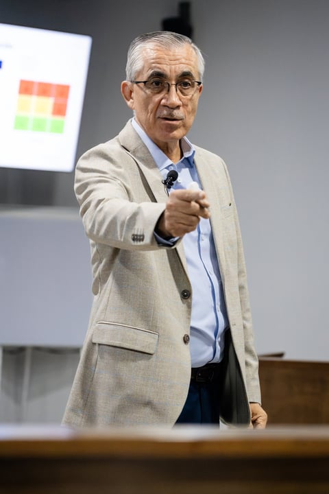 Older man with gray hair and glasses wearing a beige blazer gestures while speaking, with a colorful chart visible on a screen behind him