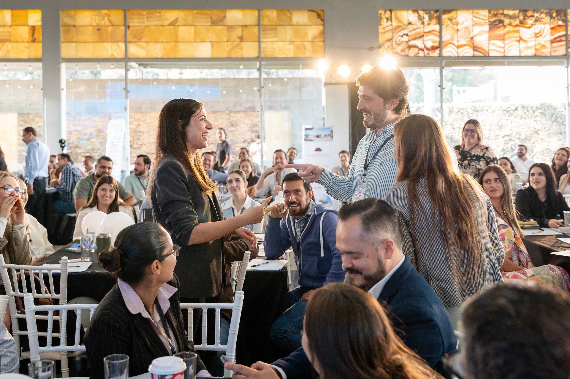 People at a conference or networking event standing and interacting in a large sunlit room with many seated attendees