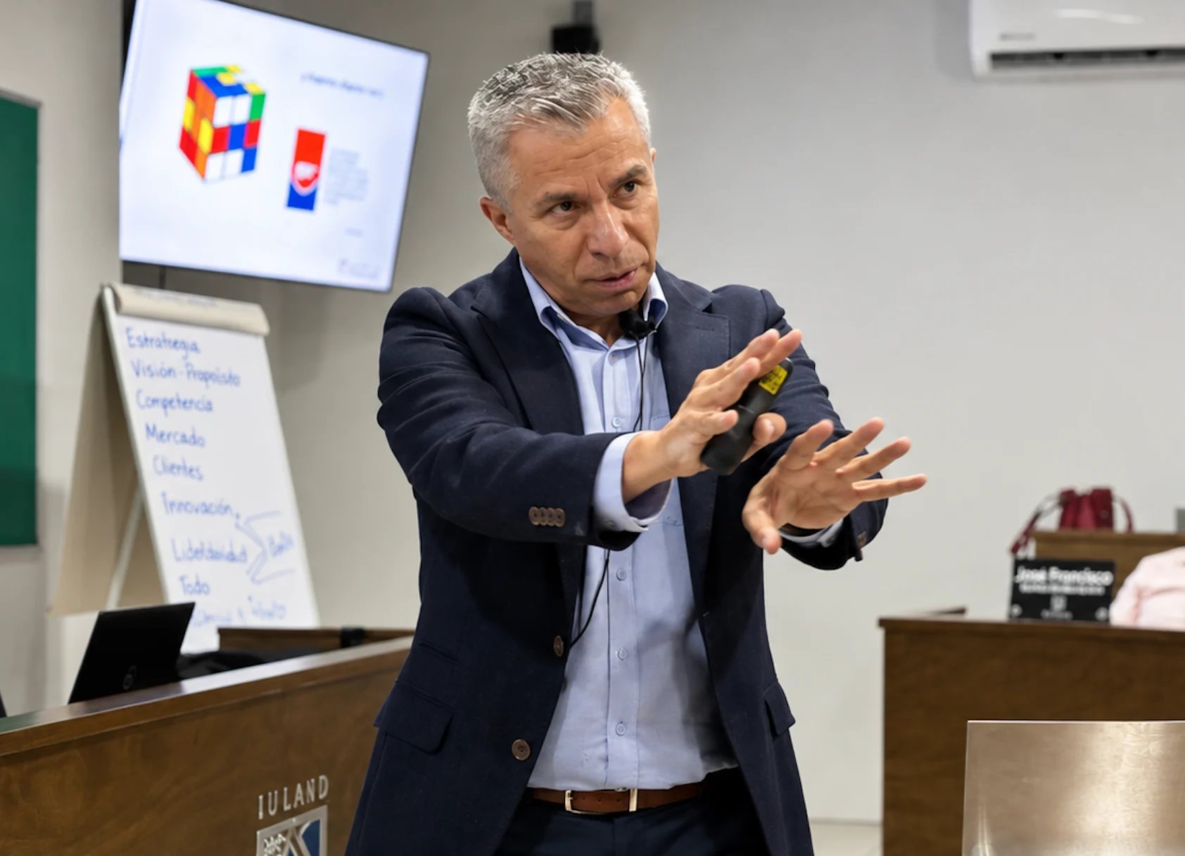 Middle-aged man in business casual attire gesturing while speaking in front of a whiteboard and projection screen during a presentation.