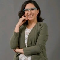 Woman wearing olive cardigan over white top, smiling and resting chin on hand against neutral gray background