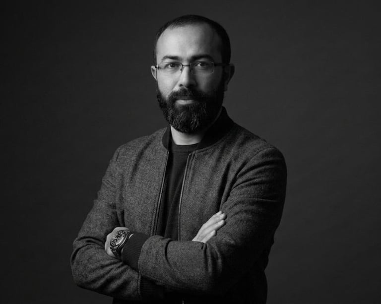 Black and white portrait of a bearded man wearing glasses and a cardigan, arms crossed against a dark background