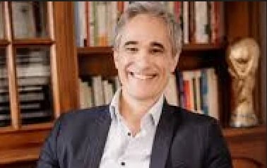 Professional man with gray hair wearing a dark blazer and white shirt, smiling in front of wooden bookshelves