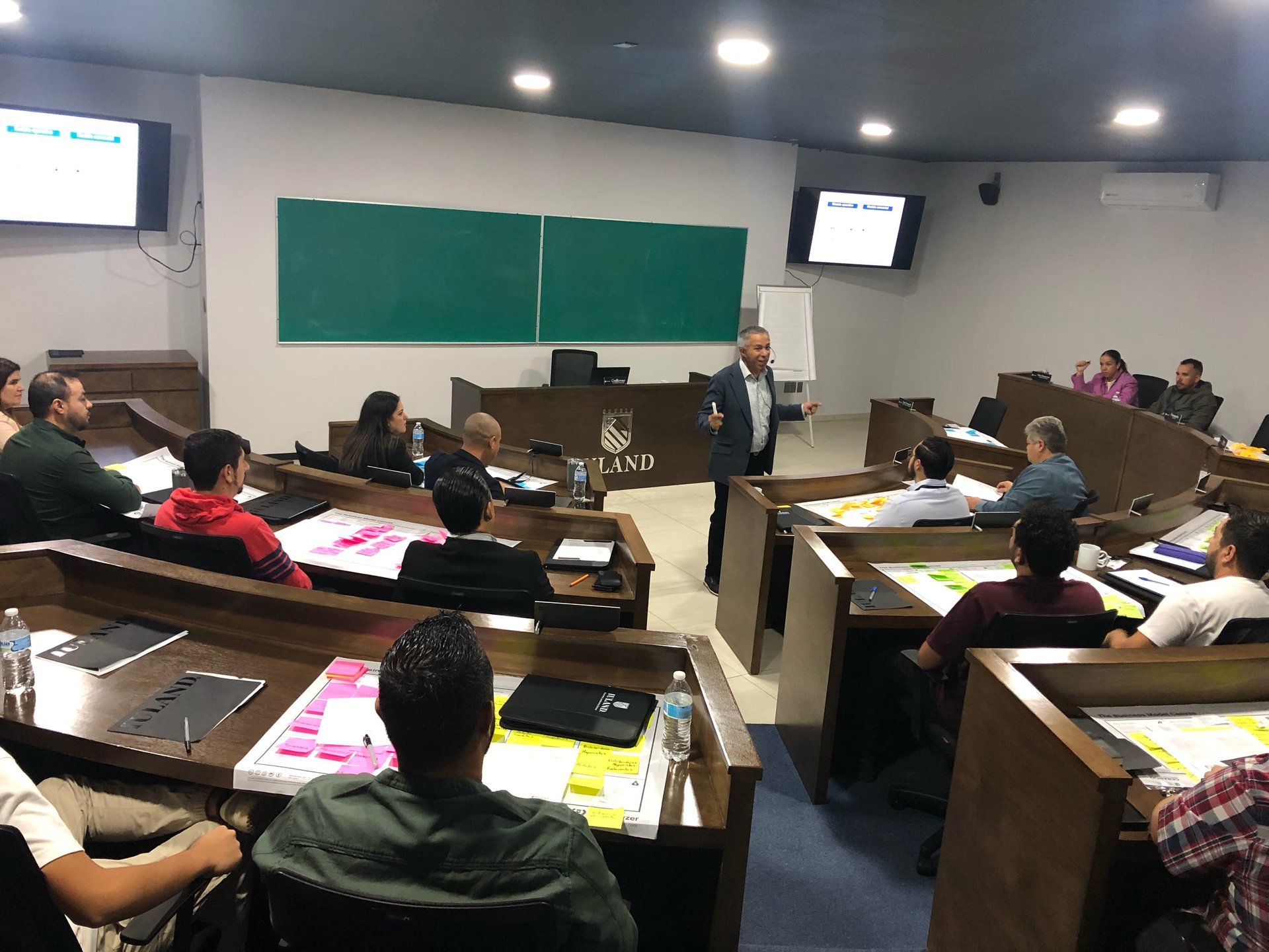 Professor teaching a university lecture hall with students seated at individual desks, green chalkboards visible at the front of the classroom.