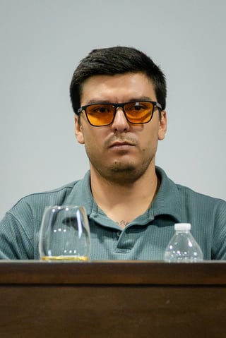 Man wearing orange-tinted sunglasses and gray polo shirt sitting at table with water bottles against white background