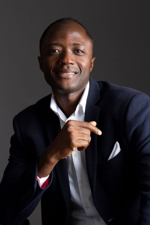 Professional headshot of a smiling man in a dark blazer and white shirt, with hand gesturing toward camera against a gray background