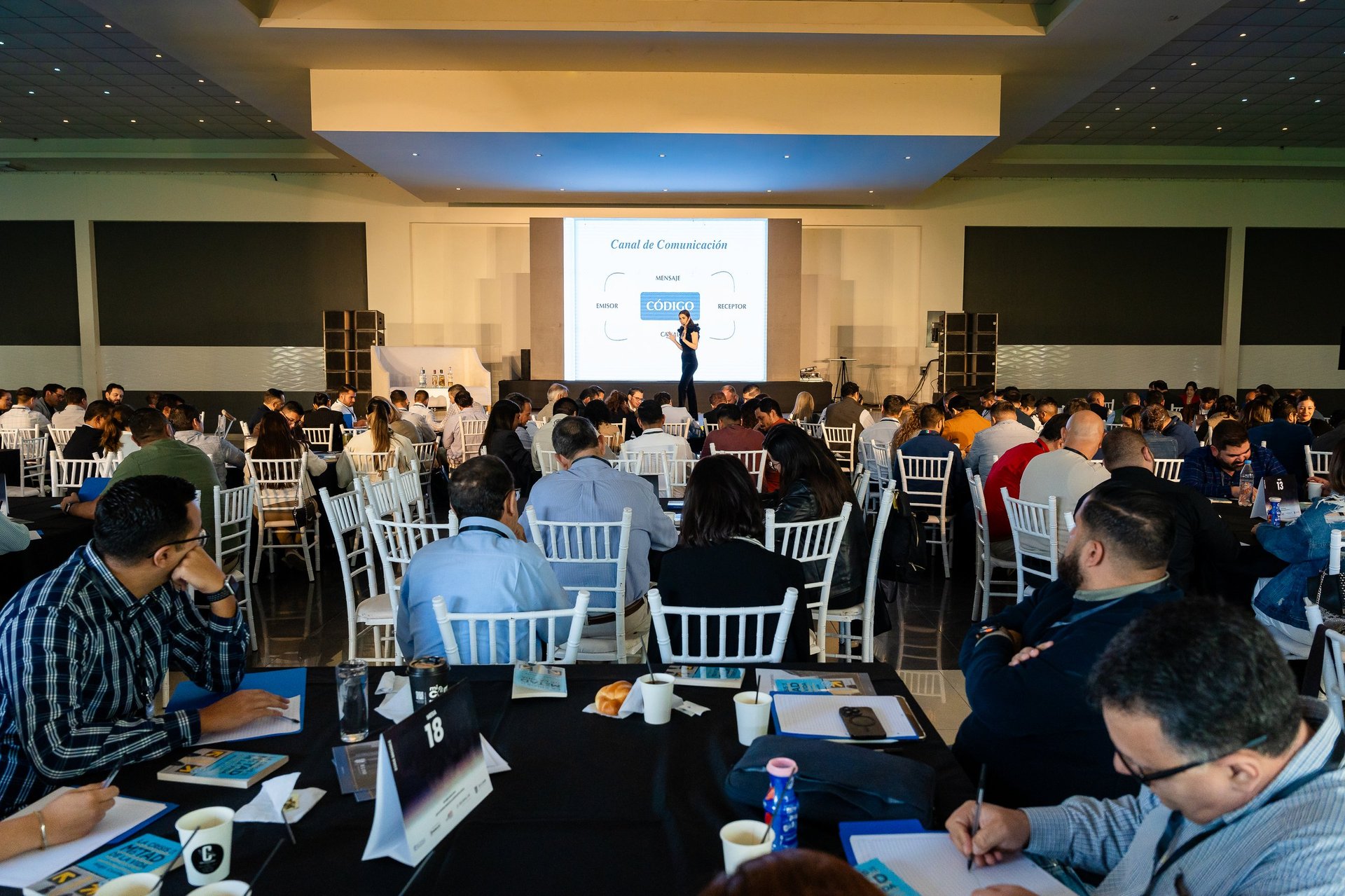 Conference hall with attendees seated at round tables facing a presenter on stage with projection screen